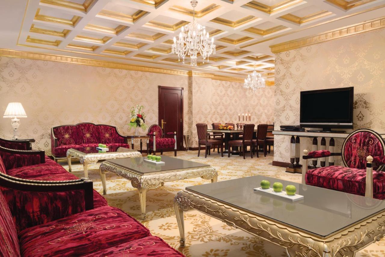 Grand Regency Doha, Trademark Collection By Wyndham מראה חיצוני תמונה The photo depicts an elegantly designed living room. The room features luxurious red sofas with ornate designs and metallic gold accents, arranged around glass-topped coffee tables that have intricate bases. A large, modern flat-screen television is 