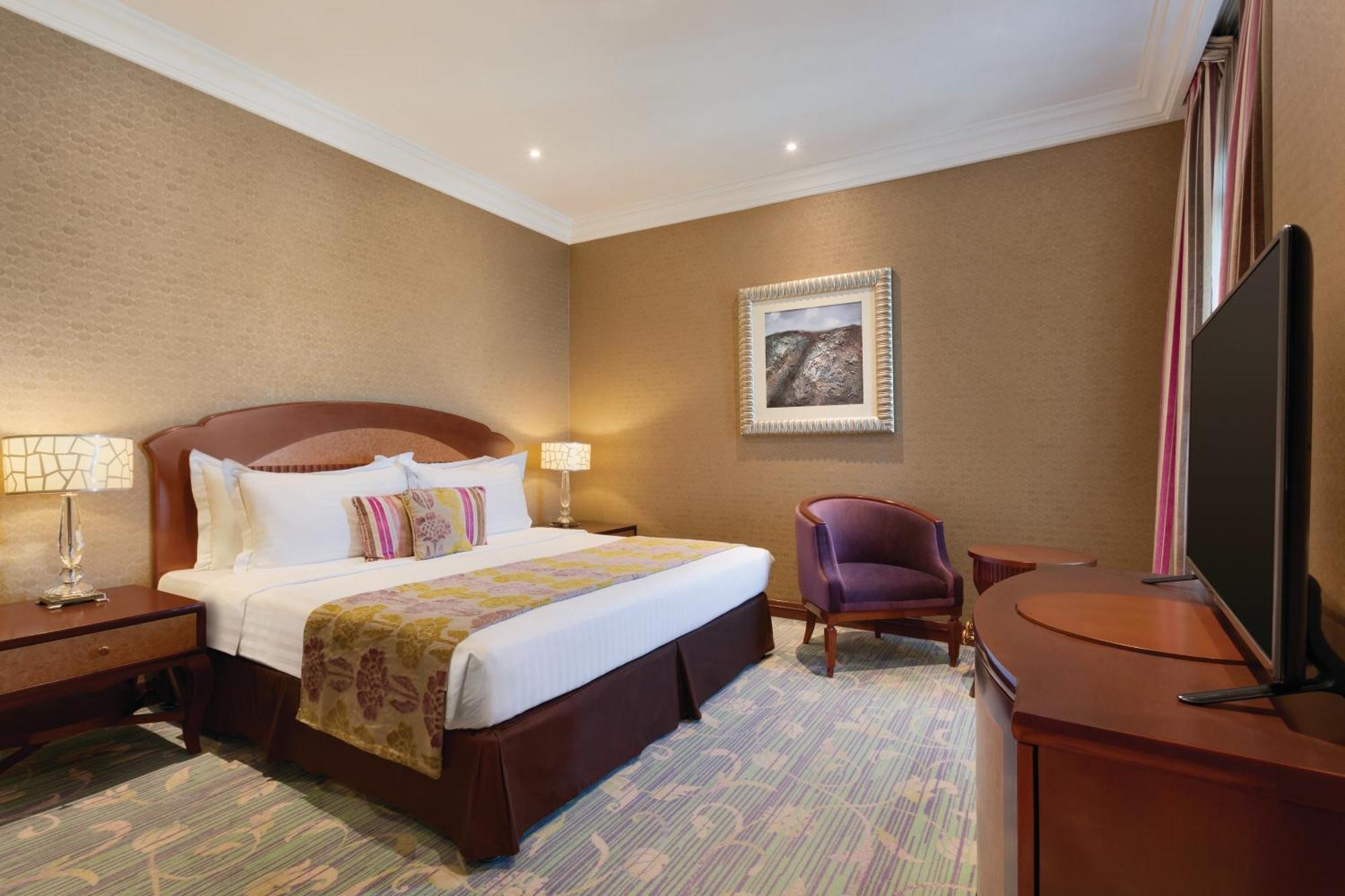 Grand Regency Doha, Trademark Collection By Wyndham מראה חיצוני תמונה The image shows a hotel room that features a cozy and elegant design. There's a large bed with white sheets and colorful decorative pillows. Next to the bed, there are nightstands with lamps. A comfortable armchair is positioned in the corner, and a 
