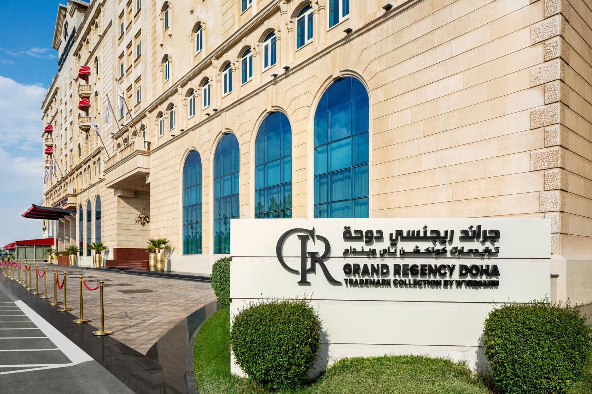 Grand Regency Doha, Trademark Collection By Wyndham מראה חיצוני תמונה The photo shows the entrance of the Grand Regency Doha, a luxury hotel. The building features a distinctive architectural style with large arched windows. There is a prominent sign featuring the hotel's name in both English and Arabic. The surroundin