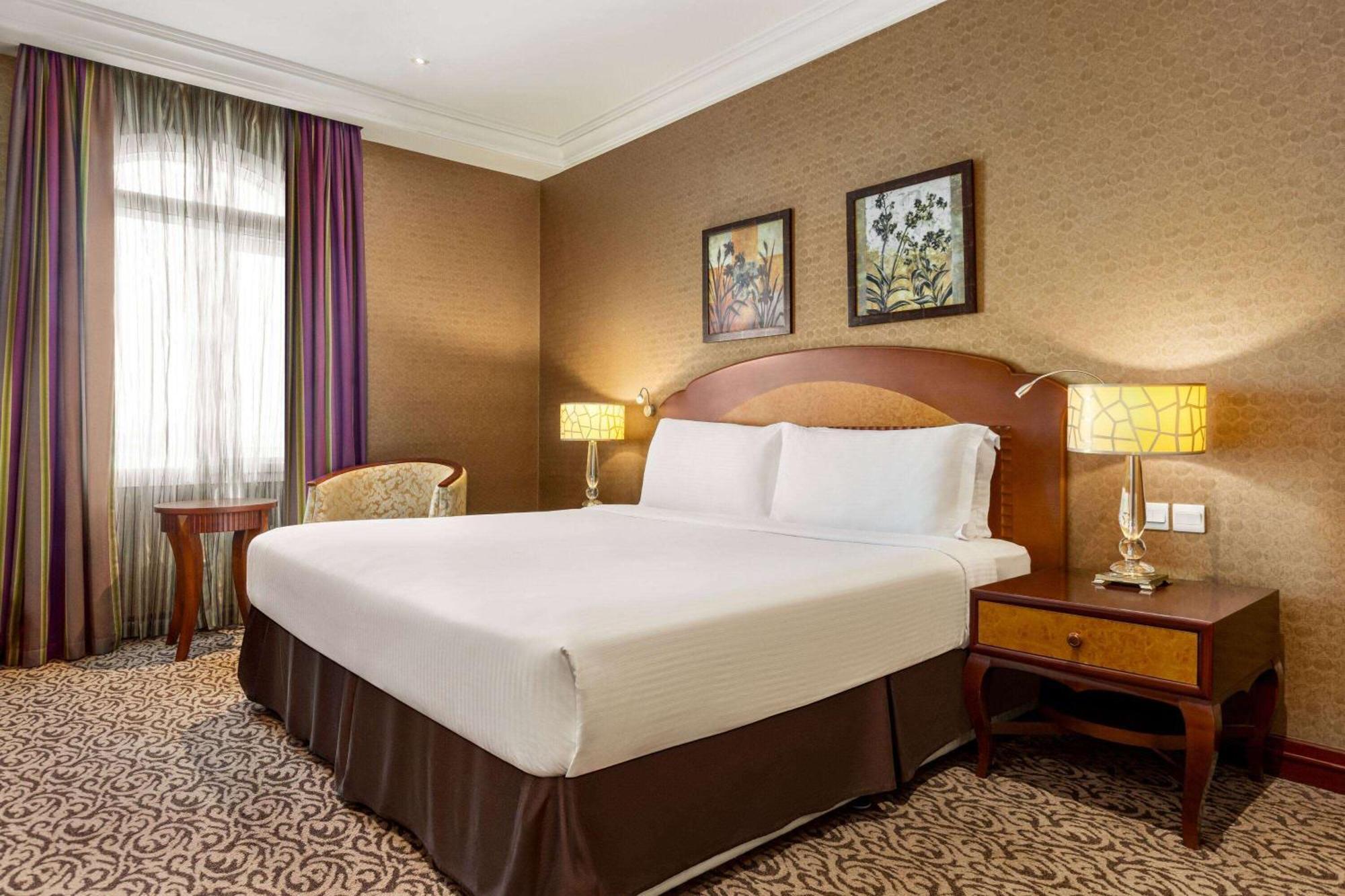 Grand Regency Doha, Trademark Collection By Wyndham מראה חיצוני תמונה The photo displays a comfortable hotel room featuring a large bed with white linens and a brown bed skirt. The room has a warm color palette, with textured brown walls and patterned carpet. There are two bedside lamps providing soft lighting next to 